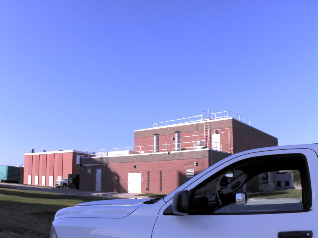 Field Surveying Services: white surveying truck in the foreground of a brick sewage treatment plant facility on a clear blue sky day